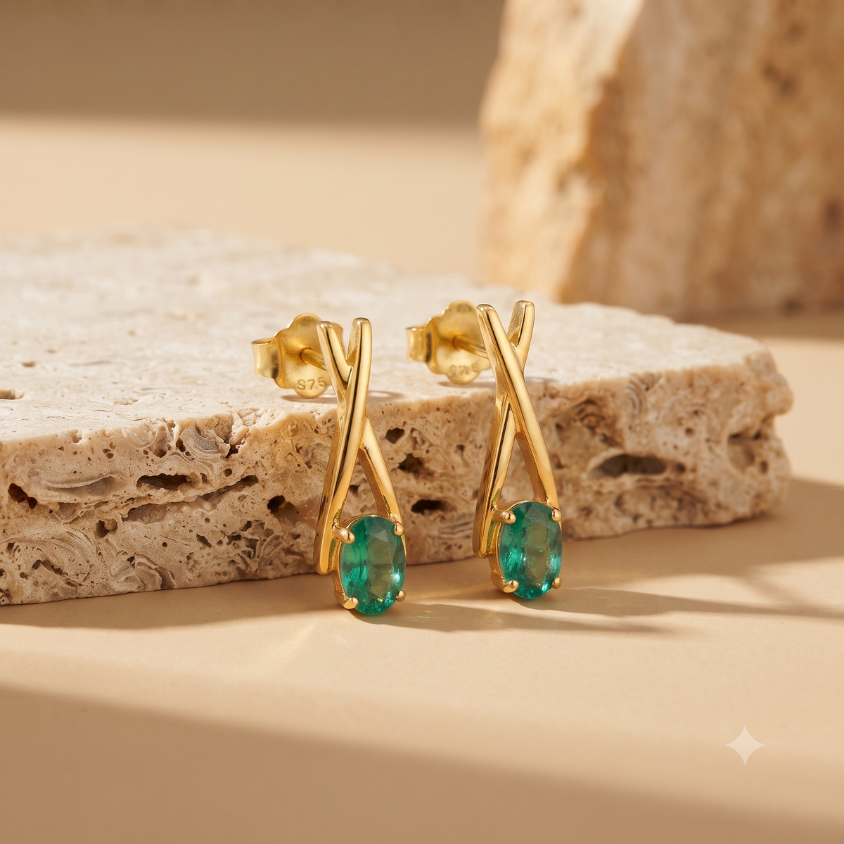 A pair of gold earrings with twisted crisscross stems and oval green gemstones, displayed on a textured beige stone slab against a warm beige background.