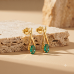 A pair of gold earrings with twisted crisscross stems and oval green gemstones, displayed on a textured beige stone slab against a warm beige background.