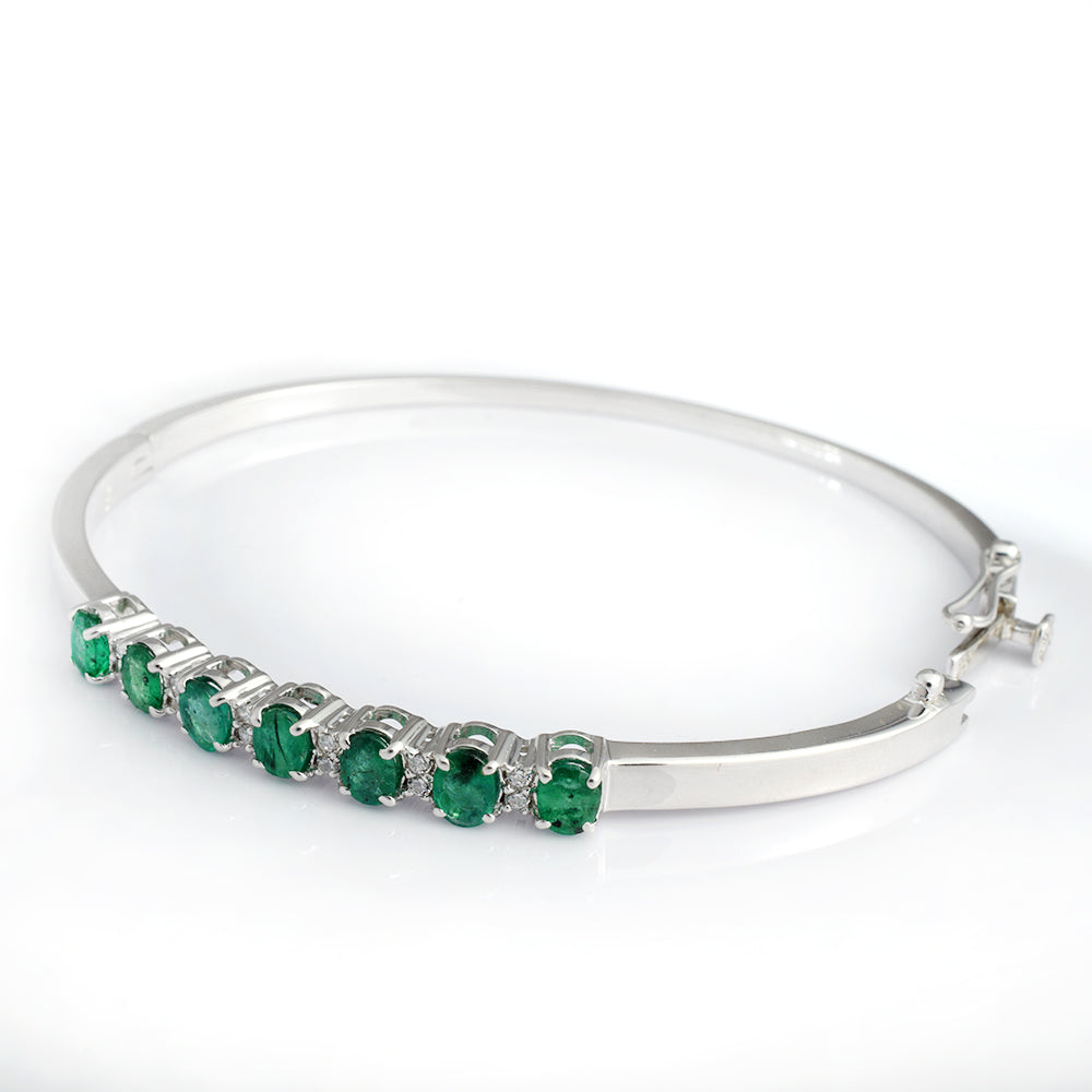 A silver bangle bracelet featuring a row of round green emerald-cut gemstones and small clear accent stones, set against a plain white background.