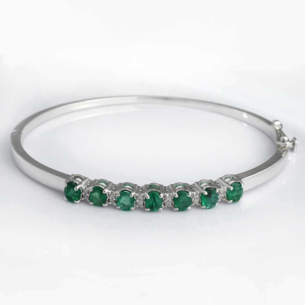 A silver-toned hinged bangle bracelet featuring a row of seven round-cut green emeralds accented by small clear stones, set against a plain white background.