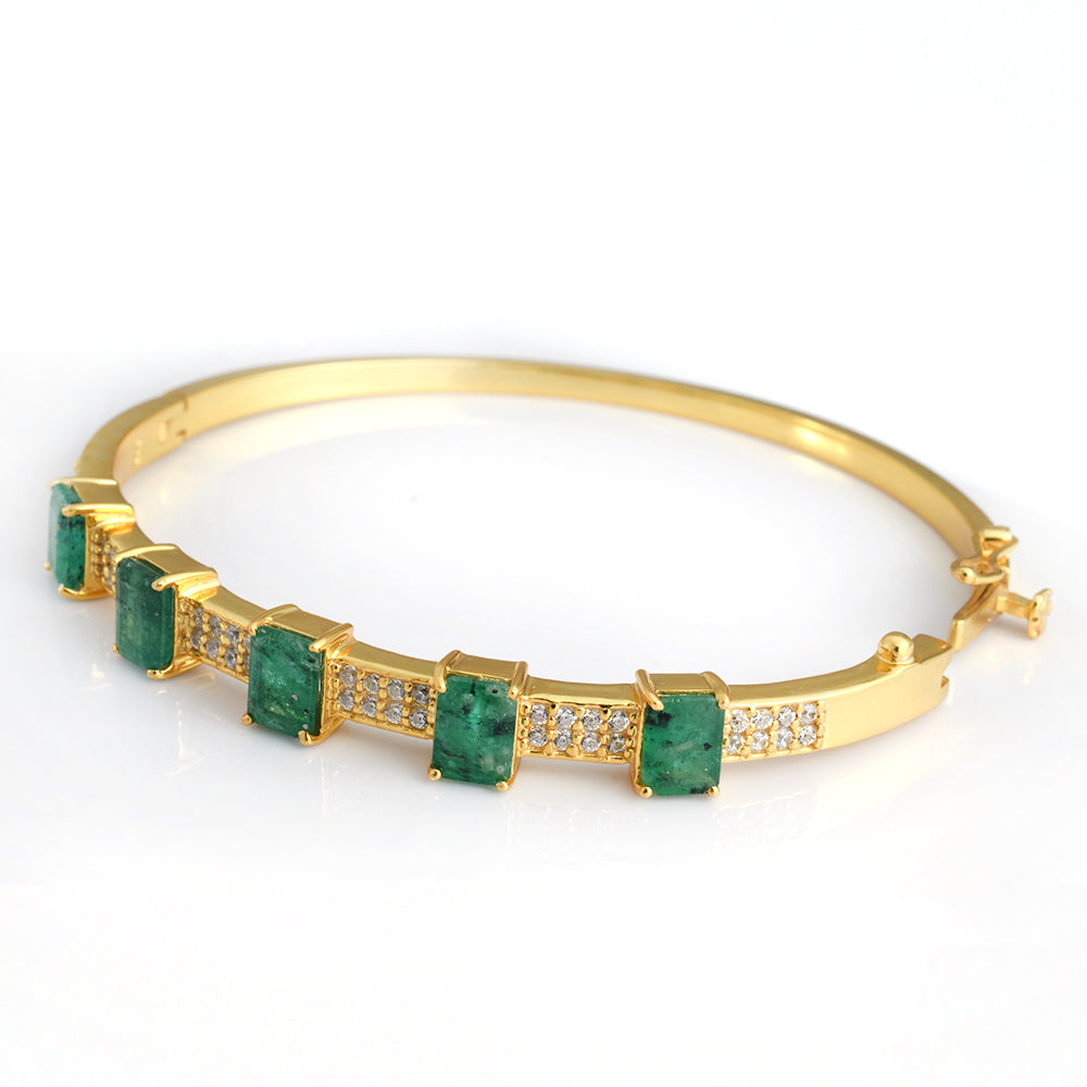 A gold bangle bracelet featuring five rectangular green emerald-cut gemstones and rows of small clear pavé stones, shown on a white background.