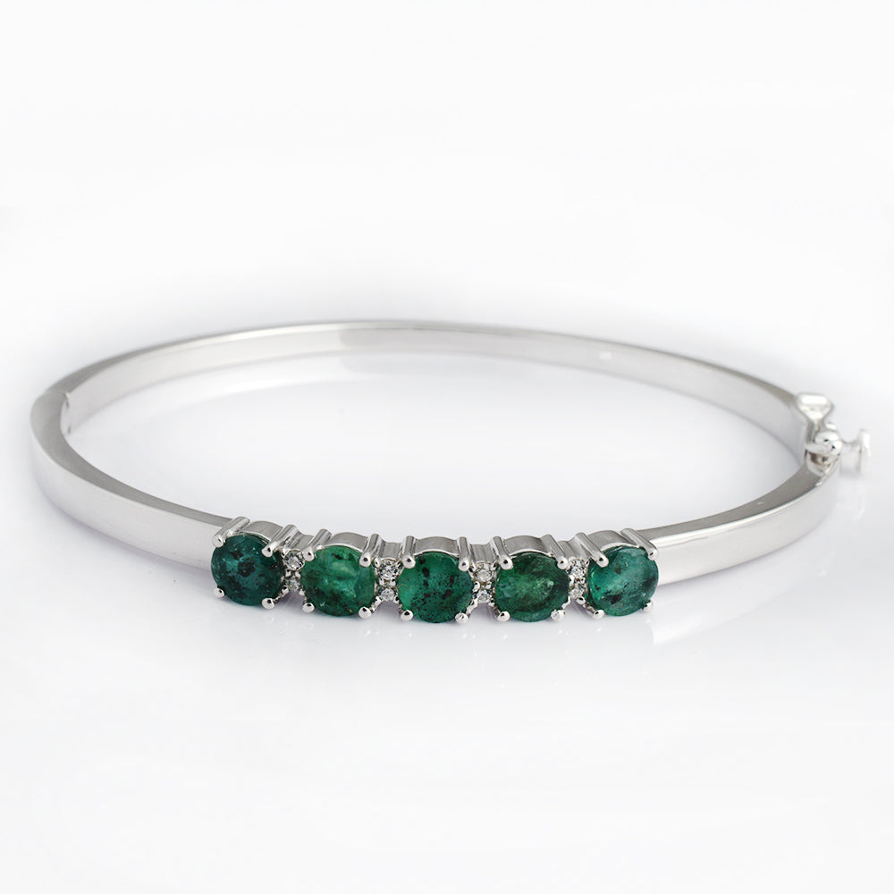 A polished silver-tone hinged bangle bracelet featuring five round-cut green emeralds set in a row along the front, displayed on a clean white background.