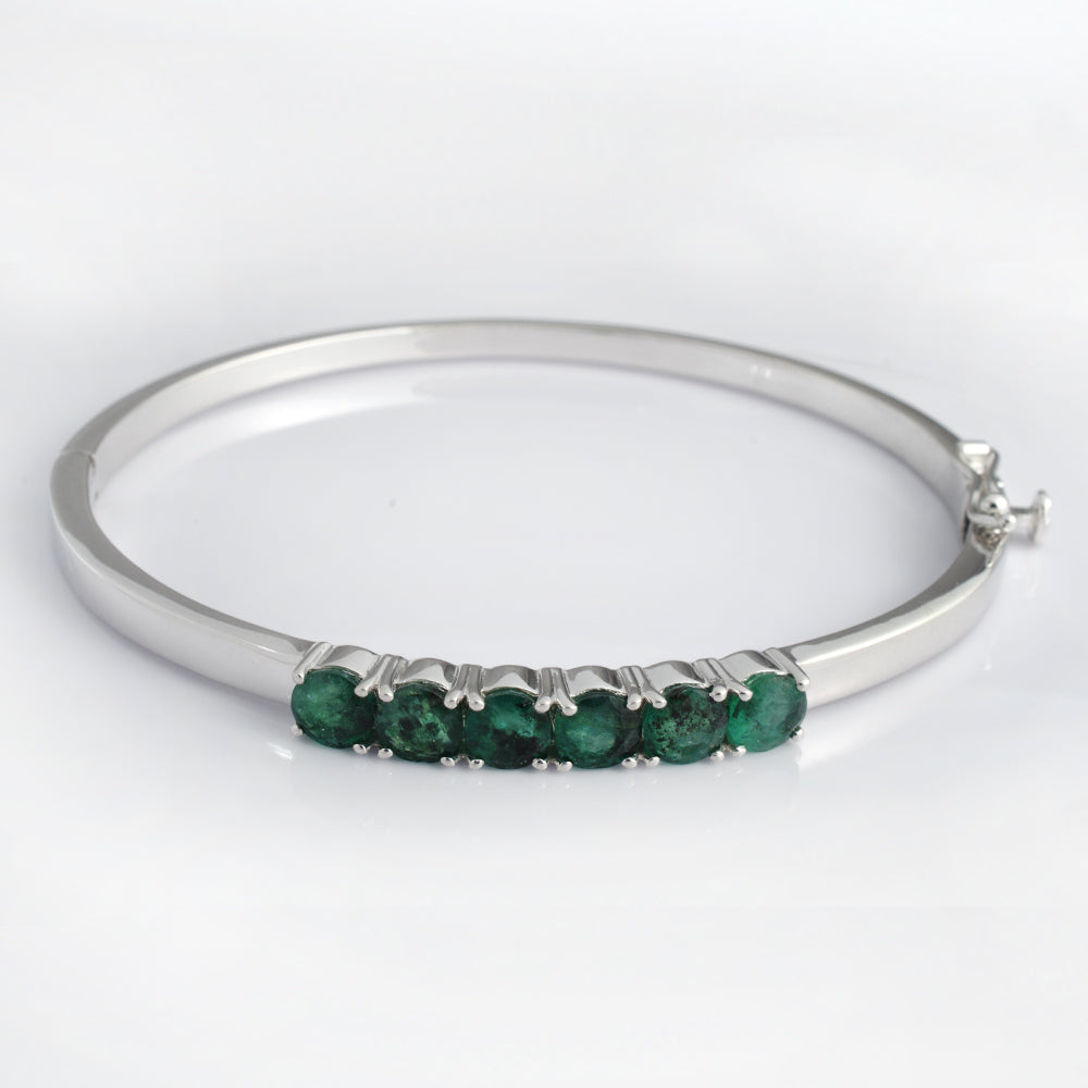 A silver bangle bracelet featuring a row of five round-cut green emeralds in a prong setting, displayed on a clean white background.