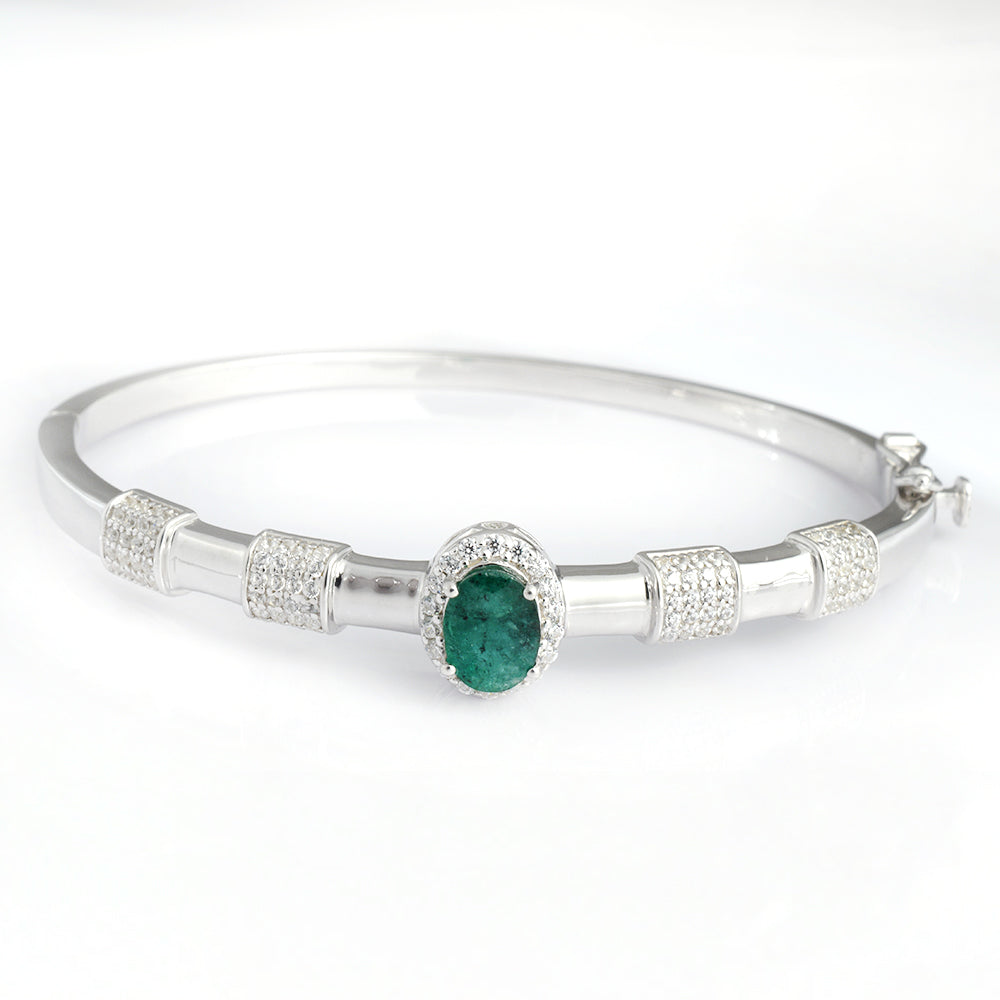 A silver-toned hinged bangle bracelet featuring a central oval green emerald stone surrounded by a halo of small clear stones, with cylindrical links embellished with pavé-set clear stones, set against a plain white background.