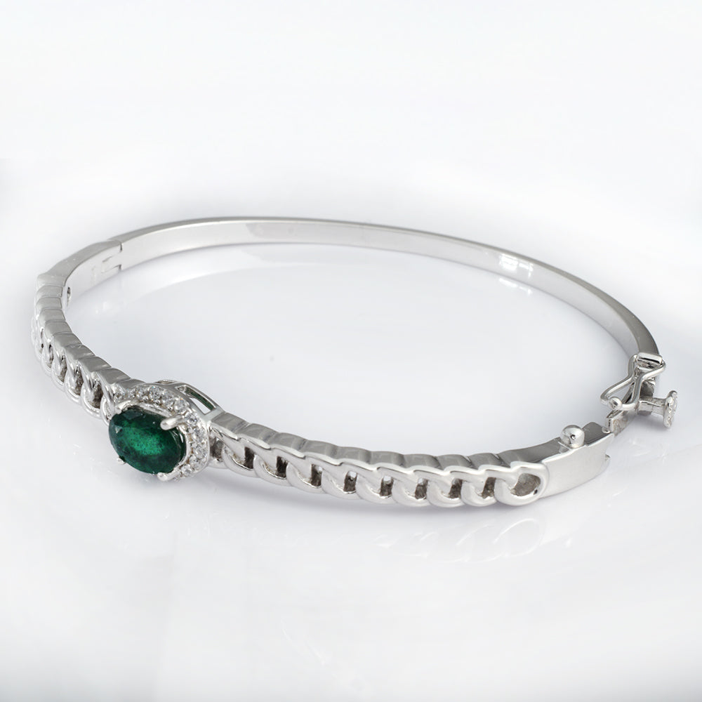 A silver bangle bracelet featuring a central oval green emerald stone surrounded by small clear stones, set on a textured silver band, shown on a white background.