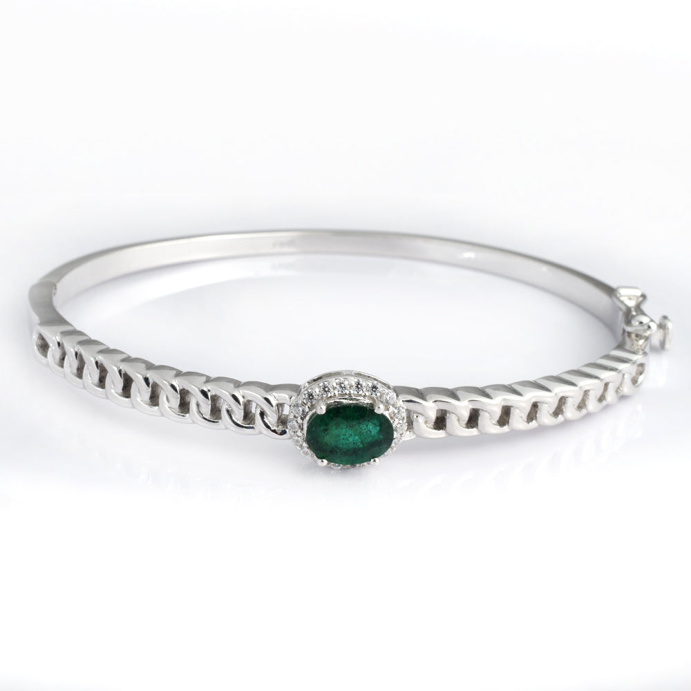 A silver bangle bracelet featuring a central oval green emerald gemstone surrounded by small clear stones, set on a polished silver chain with a textured outer band, shown on a white background.