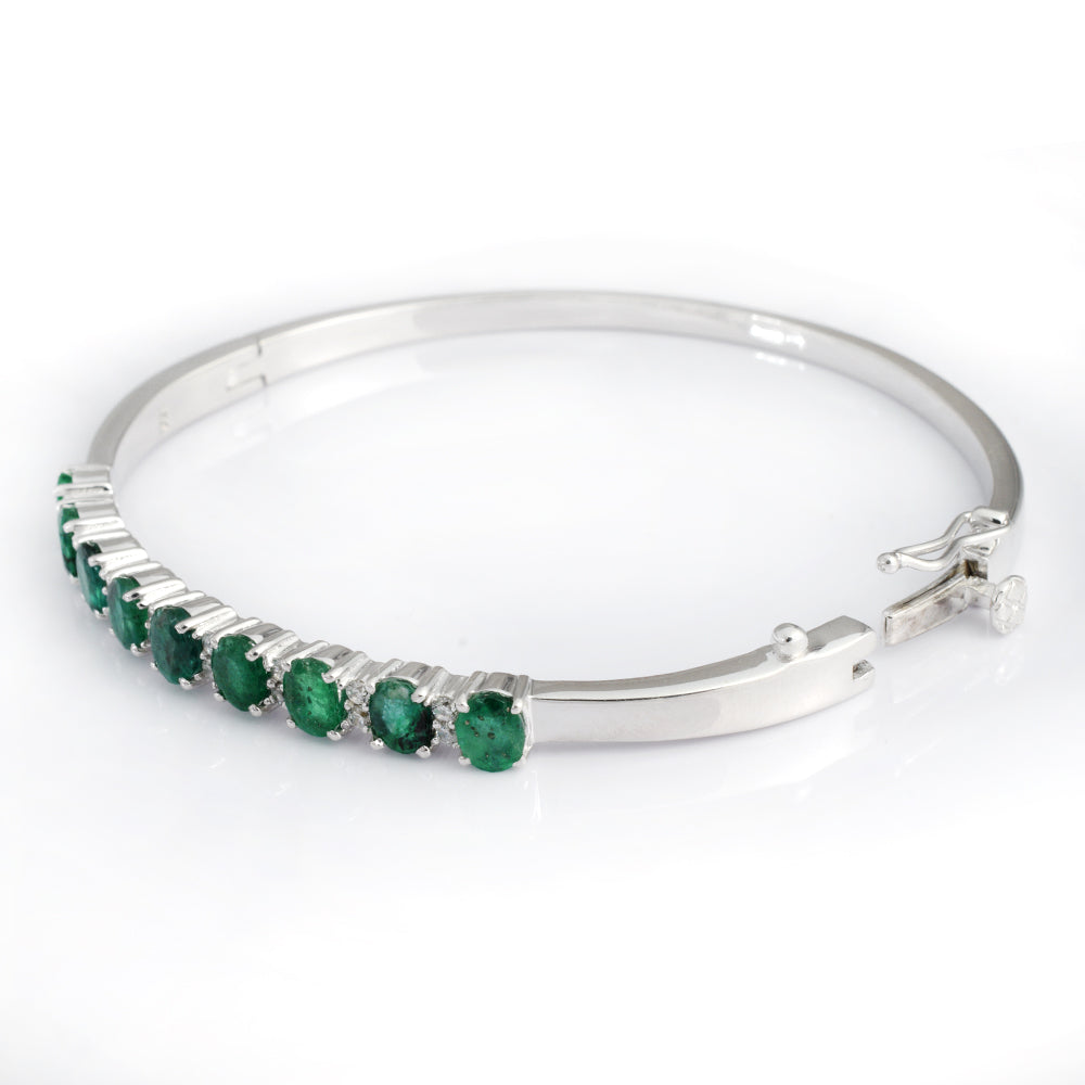 A silver-toned hinged bangle bracelet featuring a row of round green emerald-cut gemstones, set against a clean white background.