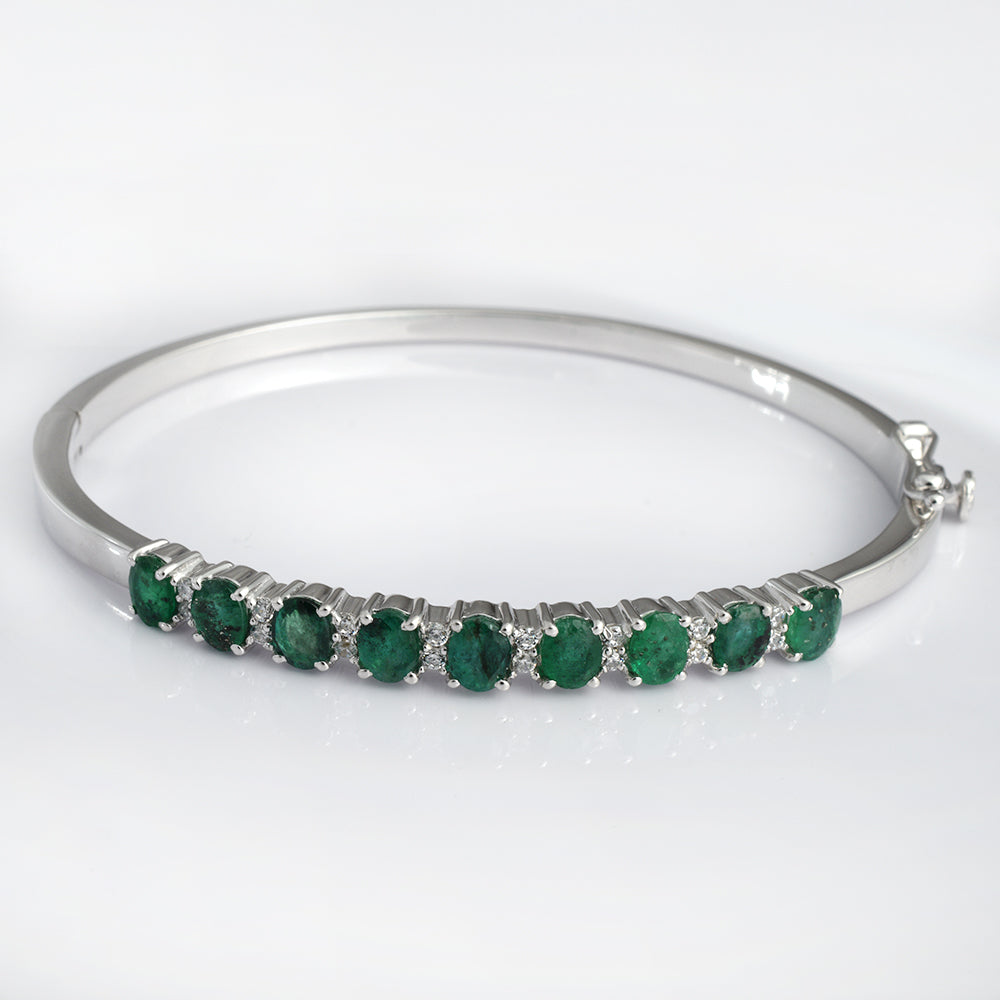 A polished silver-tone bangle bracelet featuring a row of oval-cut green emeralds in prong settings, displayed on a clean white background.