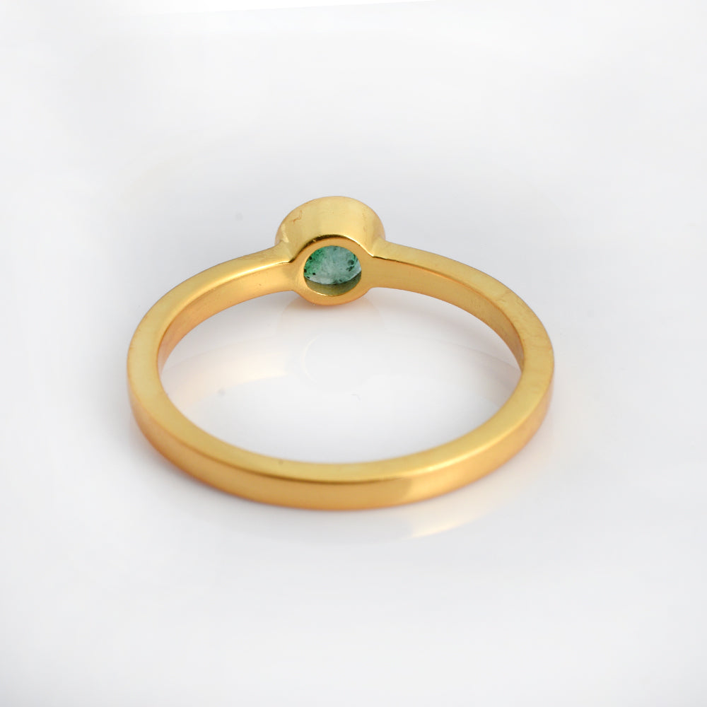 A minimalist gold ring featuring a small, round-cut green gemstone in a bezel setting, photographed on a clean white background.