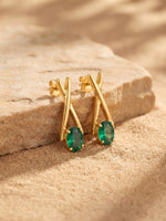 A pair of gold earrings with oval green gemstones, displayed on a textured beige stone surface with a sandy background.