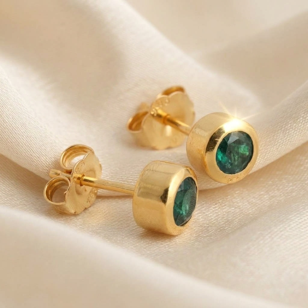 A pair of gold stud earrings with round green gemstones, displayed on a soft beige fabric background.