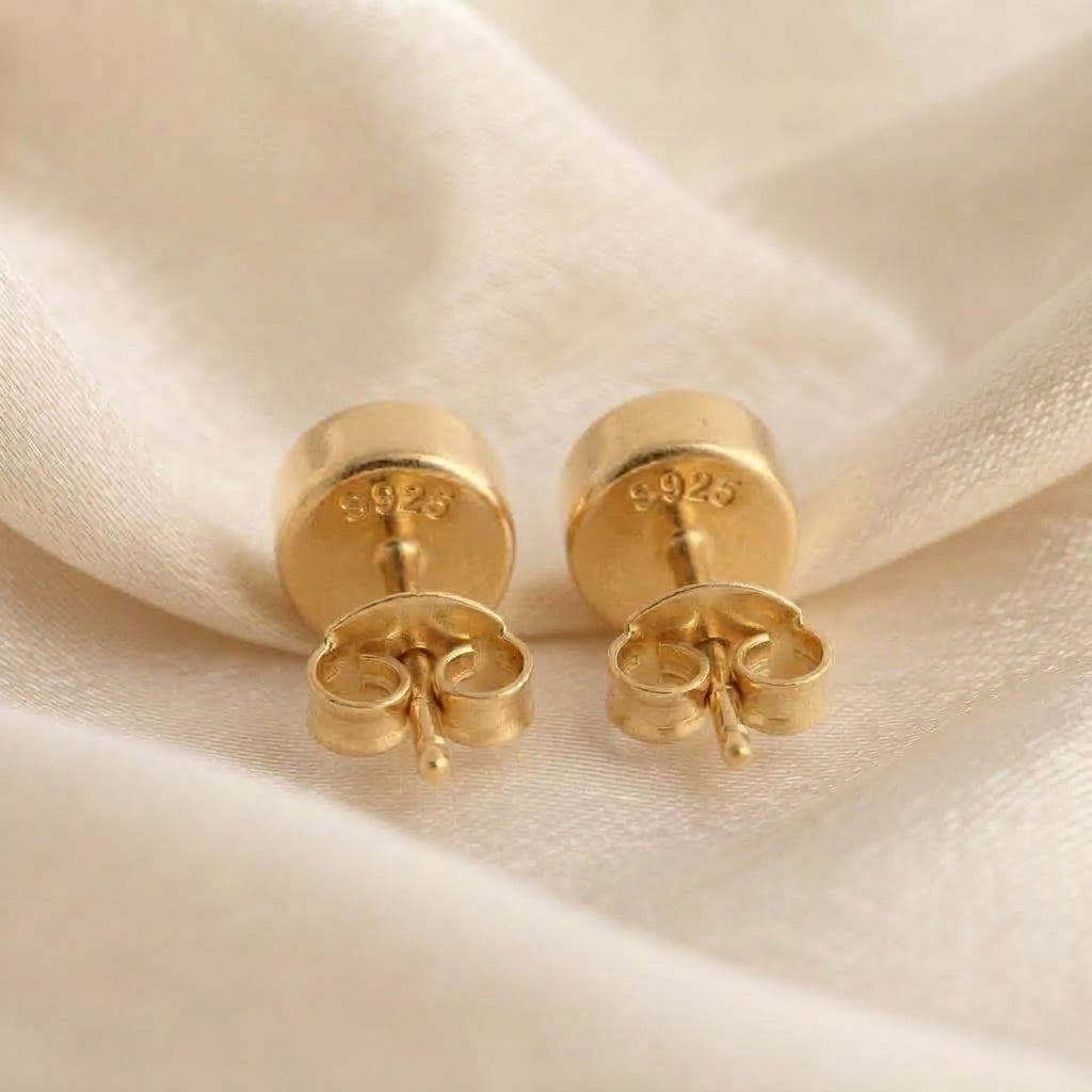 A pair of gold stud earrings with butterfly backings, displayed on a soft, draped cream-colored fabric. The back of one earring is visible, showing a '925' sterling silver purity stamp.