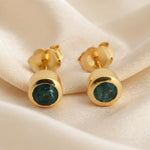 A pair of gold stud earrings with round dark green gemstone centers, displayed on a draped cream satin fabric.