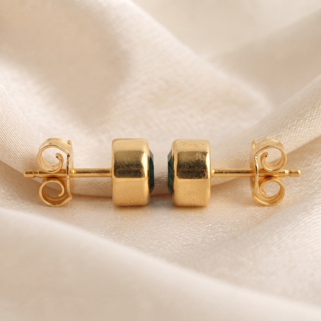 A pair of gold stud earrings with square-cut clear stones set in a dark green metal, displayed on a soft beige fabric background.