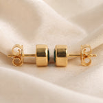 A pair of gold stud earrings with square-cut clear stones set in a dark green metal, displayed on a soft beige fabric background.