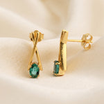 A pair of gold stud earrings with green gemstone accents, displayed on a soft beige fabric background. One earring is shown from the front with an oval-cut green stone, and the other is shown from the side with a rectangular green stone.