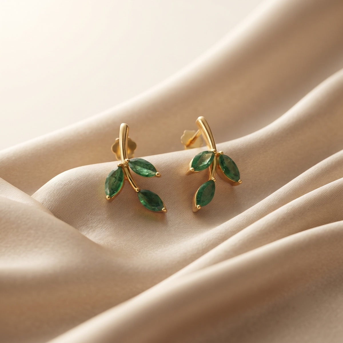 A pair of gold leaf stud earrings with marquise-cut green gemstones, displayed on a draped beige satin fabric.