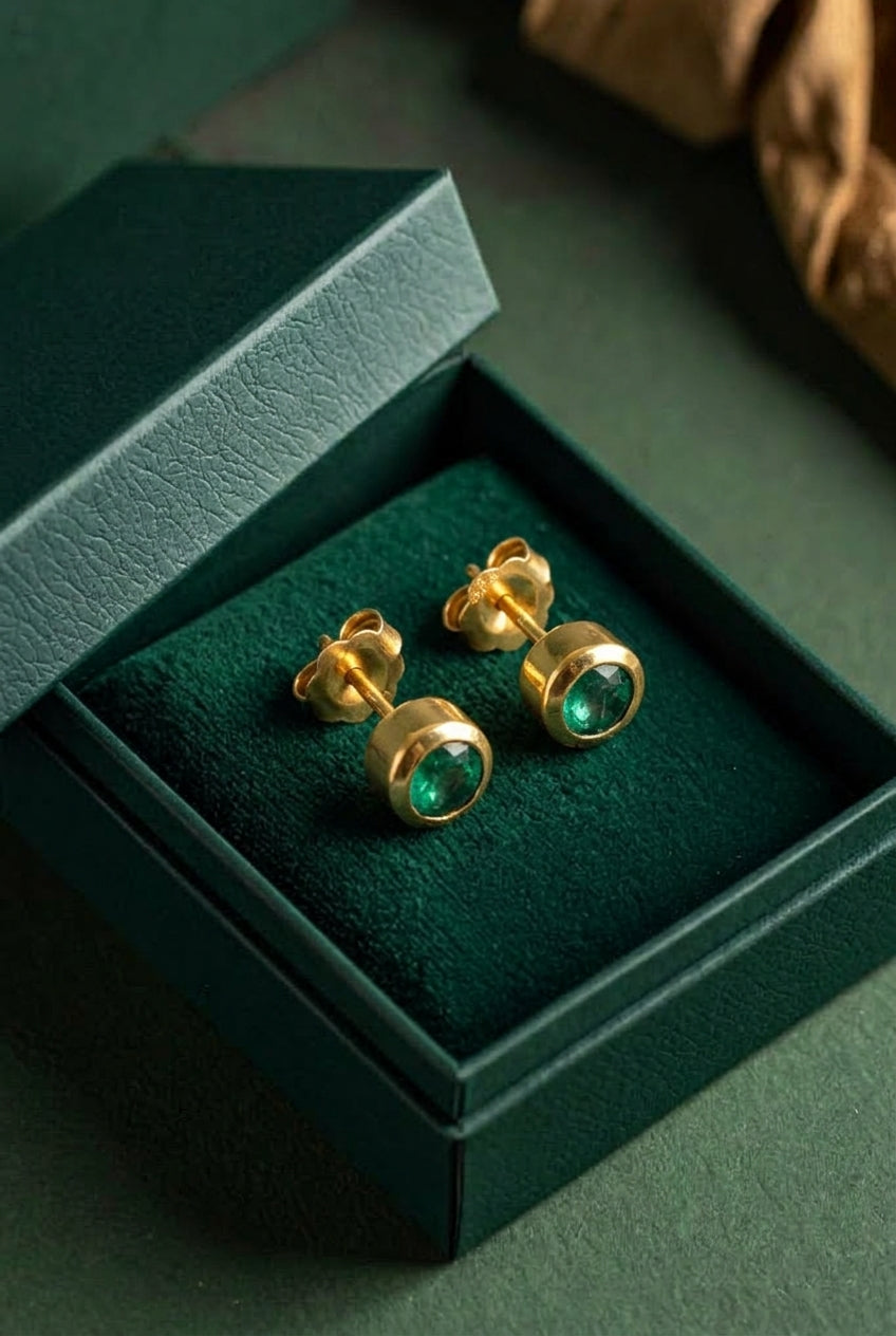 A pair of gold stud earrings with round green gemstones, presented in an open dark green jewelry box on a dark green surface.