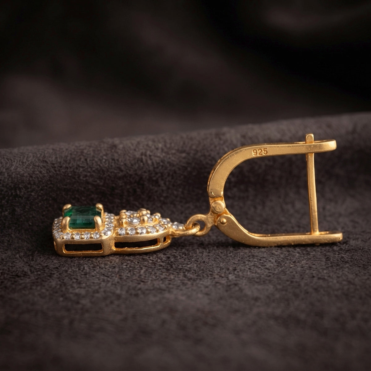 A gold earring shown in side view on a dark textured fabric background. The earring features a gold leverback clasp with a '925' sterling silver hallmark, a small gold crown detail, and a rectangular green emerald-cut gemstone surrounded by clear pavé stones.