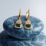 A pair of gold drop earrings with rectangular green emerald-cut center stones and small clear stones along the hangers and lower setting, displayed on stacked blue marbled stones against a light gray background.
