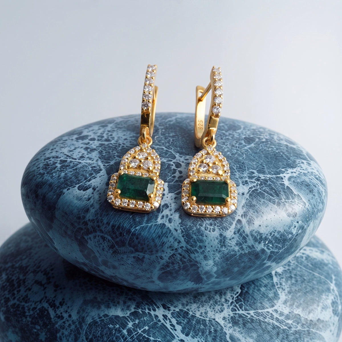 A pair of gold drop earrings with rectangular green emerald-cut center stones and small clear stones along the hangers and lower setting, displayed on stacked blue marbled stones against a light gray background.