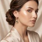 A close-up of a woman wearing a gold and green drop earring, with her hair styled in an elegant updo and a satin robe against a soft beige background.