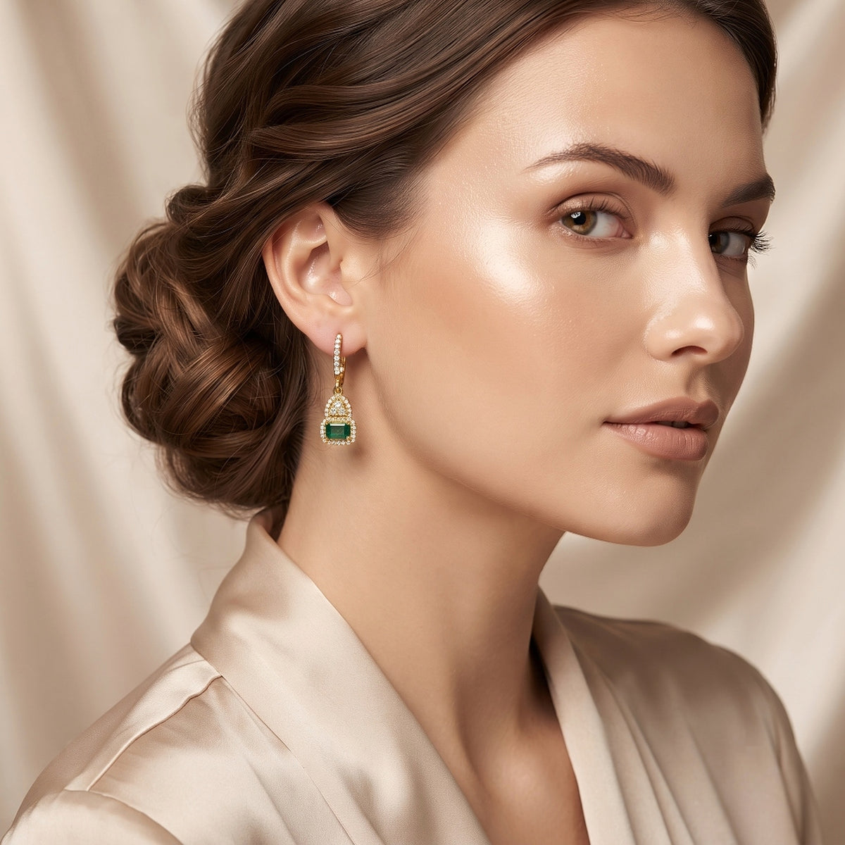 A close-up of a woman wearing a gold and green drop earring, with her hair styled in an elegant updo and a satin robe against a soft beige background.