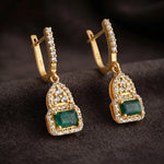 A pair of gold huggie hoop earrings with rectangular green emerald-cut stones and rows of clear pavé stones, displayed on a dark textured surface.