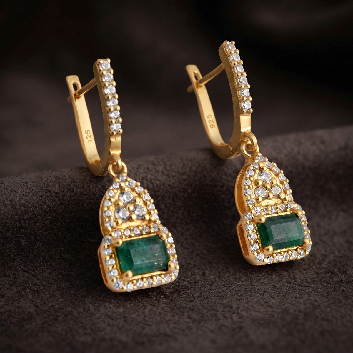 A pair of gold huggie hoop earrings with rectangular green emerald-cut stones and rows of clear pavé stones, displayed on a dark textured surface.