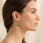 A close-up side profile of a woman wearing a delicate gold hoop earring with a small green gemstone dangle, set against a soft beige background.