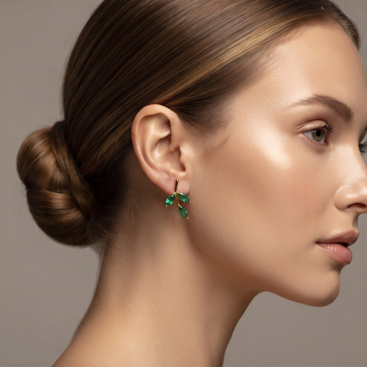 A close-up side profile of a woman with her hair in a bun, wearing a small gold hoop earring with three green marquise-cut gemstones.